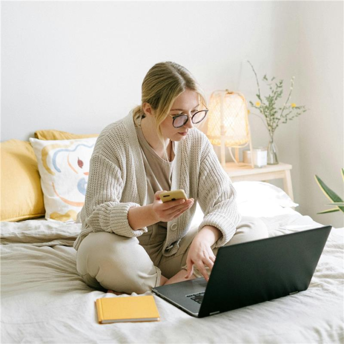 Une femme est assise sur son lit avec son ordinateur portable devant elle et son appareil téléphonique en main. Elle révise ses informations d'impôts sur les deux appareils simultanément.