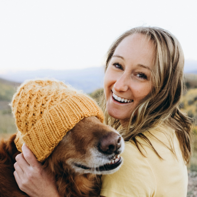 Smiling woman sitting outdoors holding a dog wearing a yellow hat
