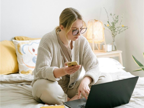 Une femme est assise sur son lit avec son ordinateur portable devant elle et son appareil téléphonique en main. Elle révise ses informations d'impôts sur les deux appareils simultanément.