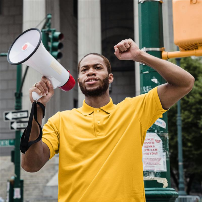 Man with a beard and wearing a yellow shirt holds a megaphone and raises one arm in the air.