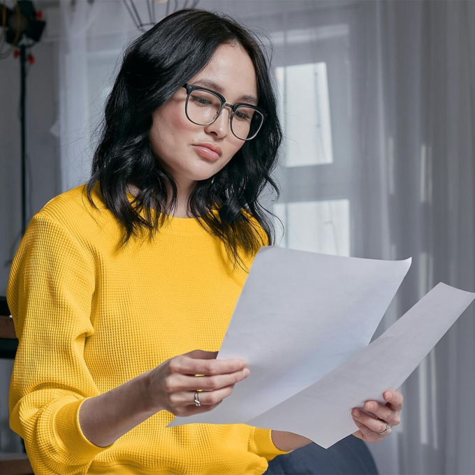A woman with glasses wearing a yellow shirt and holding pieces of paper