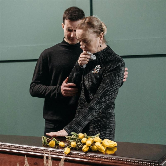 Middle-aged male and older female, both dressed in black, standing over a coffin with yellow roses laid on it.