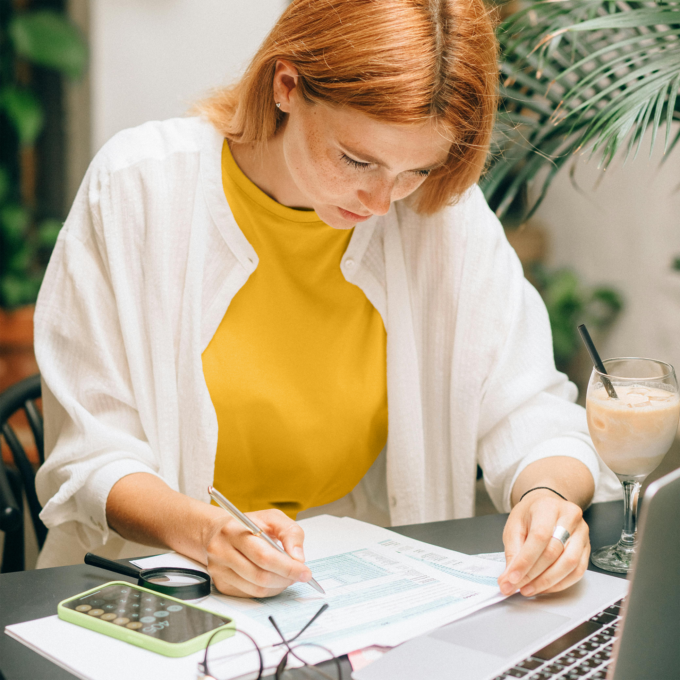 Young woman sitting at a table filling out paperwork. She has a drink next to her on the table as well as her eyeglasses and phone open to the calculator app.