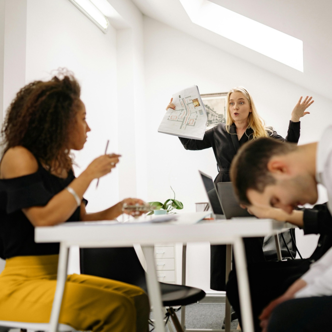 Three people in a modern office—one presenting a document, one listening, and one with head down on the table.