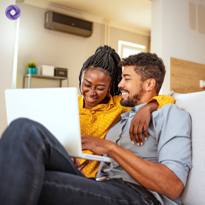 Couple sitting on the couch at home with a laptop smiling and laughing.