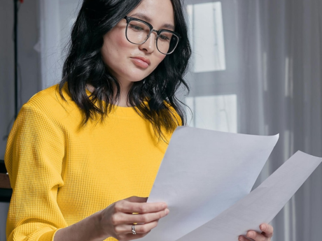 A woman with glasses wearing a yellow shirt and holding pieces of paper