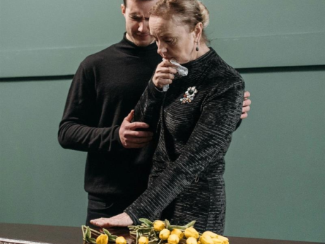 Middle-aged male and older female, both dressed in black, standing over a coffin with yellow roses laid on it.
