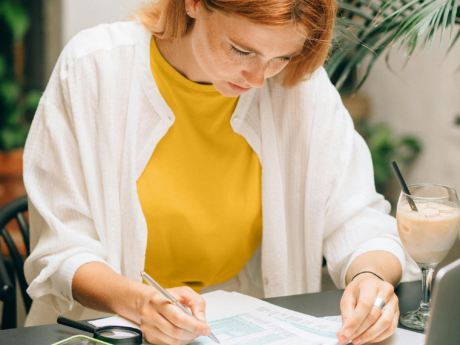 Young woman sitting at a table filling out paperwork. She has a drink next to her on the table as well as her eyeglasses and phone open to the calculator app.