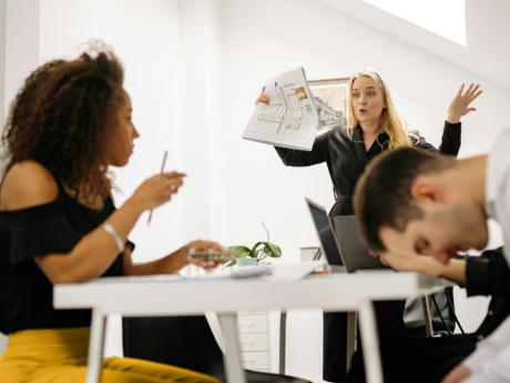 Three people in a modern office—one presenting a document, one listening, and one with head down on the table.