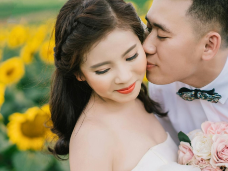 A couple embracing for a wedding photo in a sunflower field. The bride is in a white dress holding a bouquet, the groom is in a white shirt and bowtie.