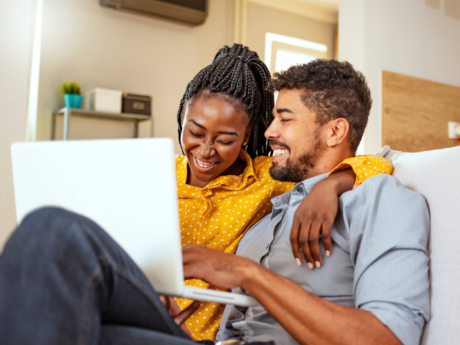 Couple sitting on the couch at home with a laptop smiling and laughing.